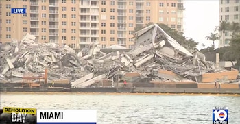 Una enorme pila de escombros de hormigón y metal de un edificio demolido se ve junto a un cuerpo de agua. Hay trabajadores y otros edificios altos en el fondo