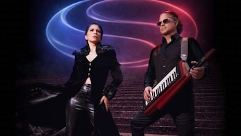 Dos personas, una mujer y un hombre, posan sobre una escalera. El hombre usa gafas de sol y sujeta un keytar. Fondo con luces de neón azules y rojas