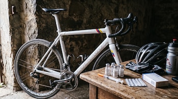 Una bicicleta blanca de carreras junto a una mesa con frascos de EPO, una jeringa, tabletas, un casco, guantes y una caramañola.