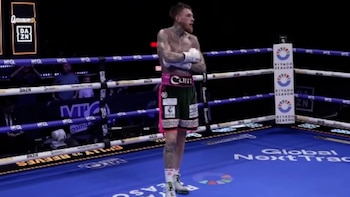 Boxeador de pie en un ring de boxeo. El boxeador tiene tatuajes, barba y viste shorts verdes y rosas, guantes y botas blancas. El ring tiene cuerdas y logos de patrocinadores