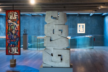 Una sala de museo con piso de madera. En el centro, una escultura abstracta de hormigón gris claro. A la izquierda, una pintura vertical colorida. Al fondo, una pared azul oscuro