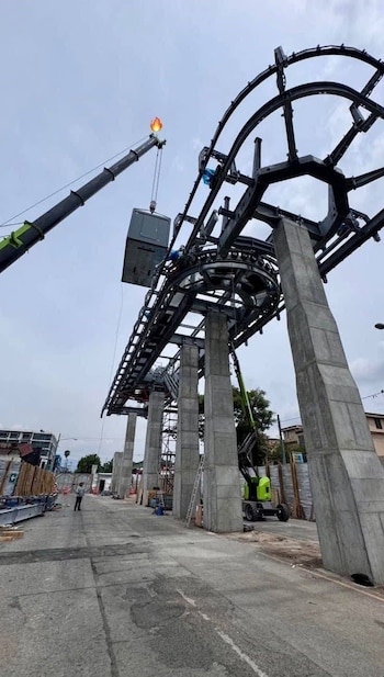 Grúa con luz en forma de llama eleva pieza metálica sobre estructura de vía elevada con pilares de hormigón en calle. Trabajador abajo. Cielo nublado
