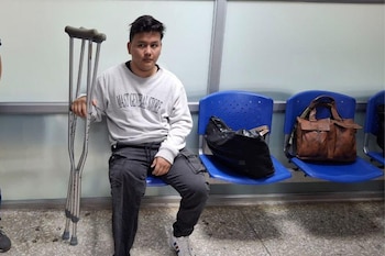 Hombre joven sentado en silla azul con muletas, sudadera gris, pantalón oscuro y zapatillas. Un bolso negro y un bolso marrón están en sillas adyacentes