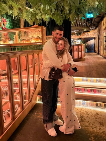 Una pareja, Karen Cavaller y Cristian Romero, se abrazan y sonríen en el centro de un pasillo de restaurante, rodeados de plantas y vitrinas iluminadas