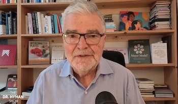 Un hombre con canas, barba y gafas se encuentra frente a un micrófono y una estantería de madera llena de libros