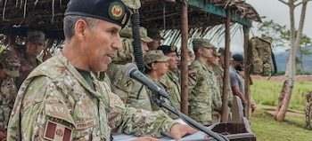 Primer plano de un oficial militar peruano en uniforme camuflado hablando en un micrófono en un podio, con otros soldados en segundo plano bajo un techo de paja