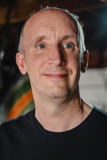 Hombre de mediana edad con cabeza calva, cabello gris en los costados, ojos azules, nariz prominente, boca cerrada en ligera sonrisa, camiseta negra