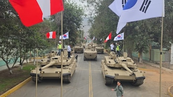 Tanques y vehículos blindados militares de color arena transitan por una carretera. Banderas de Perú y Corea del Sur ondean en postes a los lados