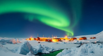 Imagen nocturna de una aurora austral verde brillante que se extiende sobre la Base Belgrano II, rodeada de nieve, témpanos de hielo y agua.