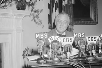 Fotografía en blanco y negro de un hombre serio, parecido a Franklin D. Roosevelt, detrás de un escritorio con numerosos micrófonos de radio de NBC, CBS y MBS