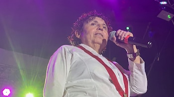 Una mujer con cabello rizado sostiene un micrófono. Viste camisa blanca y tirantes rojos. La escena tiene luces de escenario violetas y verdes.
