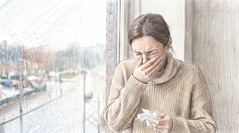 Una mujer con suéter beige se cubre la boca con la mano, sostiene un pañuelo de papel y mira a través de una ventana con gotas de lluvia.