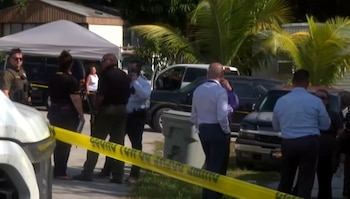 Agentes de la ley y civiles en una escena al aire libre con cinta amarilla, autos, camionetas, una tienda blanca, casas, palmeras y un cesto de basura