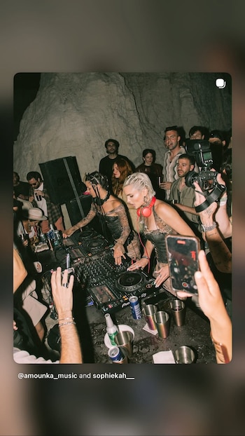 Dos mujeres con auriculares pinchan música en una consola DJ. Varias personas observan, algunas con móviles. Hay bebidas en una mesa y una pared de roca