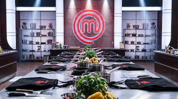 Cocina profesional con una larga mesa central equipada con mandiles negros, cacerolas, cuchillos e ingredientes frescos, y el logo de MasterChef México en el fondo.