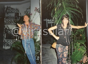 Indio Solari y Virginia, cubiertos de serpentinas, posan al aire libre. Él lleva camisa de colores y jeans; ella, blusa oscura y pantalones estampados, con plantas tropicales