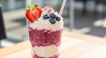 Vaso de Frappuccino de frutos rojos con capas de batido morado y crema blanca. Decorado con crema batida, arándanos, media fresa y un popote rayado.