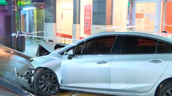 Primer plano de un coche plateado chocado de frente contra una estructura metálica en la calle, con cinta de precaución roja y blanca visible