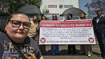 Alejandra Ley, de gafas, toma una selfie. Detrás, seis personas sostienen una pancarta blanca y roja frente al edificio del Gobierno de México y Centro Federal de Conciliación
