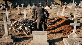 Sepultureros de espaldas cavan en un cementerio rodeado de cruces de piedra, palas, flores y una lápida con la inscripción Familia López.