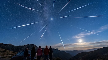 Vista nocturna de un grupo de personas observando múltiples meteoros cruzar un cielo estrellado sobre montañas, con una luna brillante y un coche.