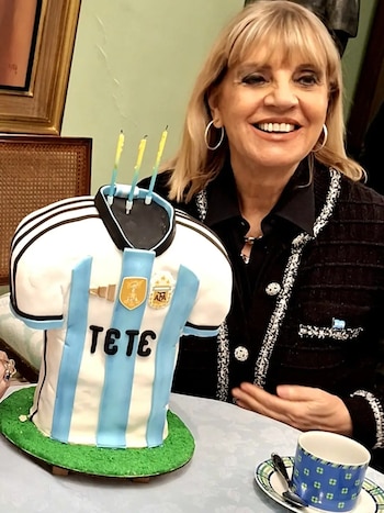 Retrato de mujer sonriente junto a pastel con forma de camiseta de fútbol argentina, con el nombre Teté, y tres velas encendidas. Taza y plato sobre mesa