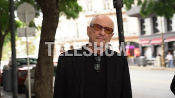 Primer plano de un hombre calvo con gafas de sol anaranjadas y chaqueta negra, de pie en una calle urbana con árboles