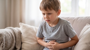 Niño sentado en un sofá con las manos sobre el abdomen. Viste camiseta gris y está frente a una ventana borrosa. Hay una manta a cuadros en el sofá.