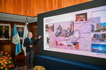 Hombre con barba apunta a una pantalla grande que muestra un diagrama del proyecto Metro Riel, con un mapa de ruta, estaciones, edificios y banderas de Guatemala al fondo