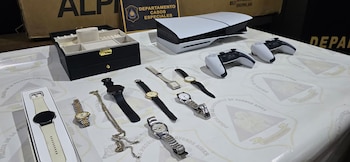Objetos incautados por la policía: una consola PlayStation 5, dos mandos, una caja de joyas, siete relojes de pulsera y una cadena dorada sobre una mesa