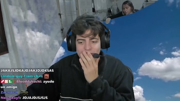 Un joven con auriculares se cubre la boca frente a la cámara mientras un fondo azul de cielo deja al descubierto una cocina con una mujer al fondo