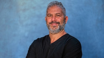 Un hombre con cabello gris, barba, bigote y camisa negra mira al frente contra un fondo azul.