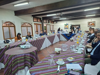 Grupo de personas sentadas en mesas cubiertas con manteles estampados en un salón. Se observan bebidas, laptops, cuadernos y un proyector