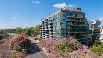 Vista aérea de un moderno edificio de apartamentos con balcones de vidrio y una calle flanqueada por árboles con flores rosadas bajo un cielo azul claro