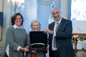 Tres personas sonríen: una mujer mayor de cabello gris en el centro, flanqueada por una mujer de cabello oscuro y un hombre calvo, sosteniendo una caja con una placa