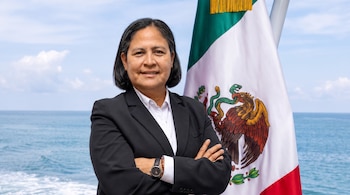 Una mujer de cabello oscuro con traje negro y camisa blanca sonríe, brazos cruzados, frente a la bandera de México con el mar y el cielo de fondo.
