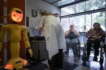 Robot amarillo con pantalla que muestra un rostro, frente a una médica con bata y dos mujeres mayores en sillas de ruedas