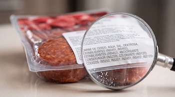 Un paquete transparente con carne procesada y una lupa sobre la etiqueta que muestra ingredientes y códigos de aditivos INS.