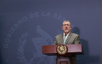Primer plano de un hombre maduro con traje gris, camisa azul claro y corbata, gafas y barba, hablando en un podio de madera con un escudo dorado