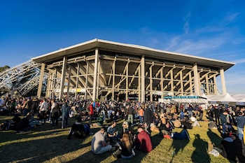 Vista panorámica de una multitud de personas sentadas y de pie sobre césped verde, frente a un gran estadio de concreto con cielo azul claro