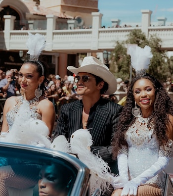 Bruno Mars sonríe en un coche descapotable, lleva sombrero vaquero blanco y gafas de sol, junto a dos mujeres con trajes brillantes y tocados de plumas