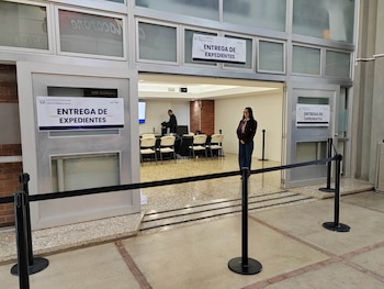 Una puerta de cristal con letreros de entrega de expedientes, barreras de cinta negra y dos personas en una sala con sillas