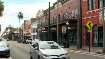 Vista frontal de una calle principal con coches aparcados y en movimiento, edificios de ladrillo de dos pisos, y palmeras bajo un cielo nublado