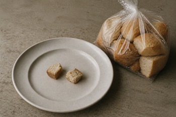 Un plato cerámico con dos pequeños trozos de pan junto a una bolsa transparente con pan troceado sobre una superficie gris.
