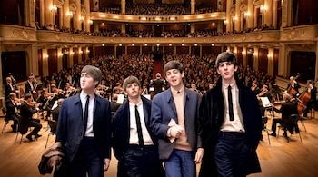 Los cuatro integrantes de The Beatles están en primer plano frente a una orquesta y un público reunido en las gradas de un teatro.