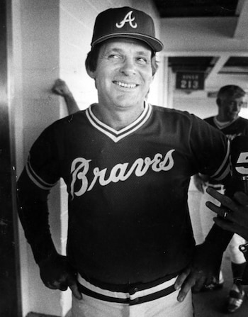 Retrato en blanco y negro de Bobby Cox sonriendo, de pie con las manos en las caderas, vistiendo el uniforme de béisbol de los Atlanta Braves y una gorra con la 'A'