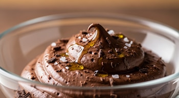 Primer plano de mousse de chocolate oscuro en un cuenco de vidrio, con textura aireada, rociada con aceite de oliva, escamas de sal y virutas de chocolate.