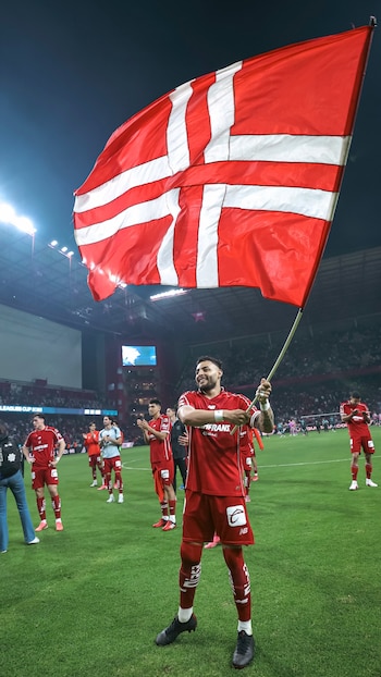 Jugador de fútbol con uniforme rojo sostiene una bandera rojiblanca con cruz escandinava en un estadio, otros jugadores y público visible