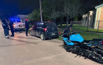 Una motocicleta celeste estacionada, un automóvil negro detenido y personas junto a una camioneta patrullera en una calle de noche