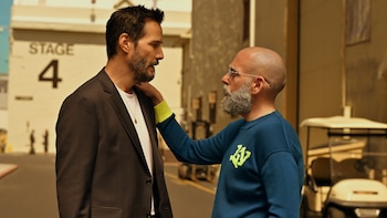 Keanu Reeves, con barba y traje oscuro, mira a Jonah Hill, calvo con barba gris y suéter azul, en un exterior con el cartel 'STAGE 4' visible
